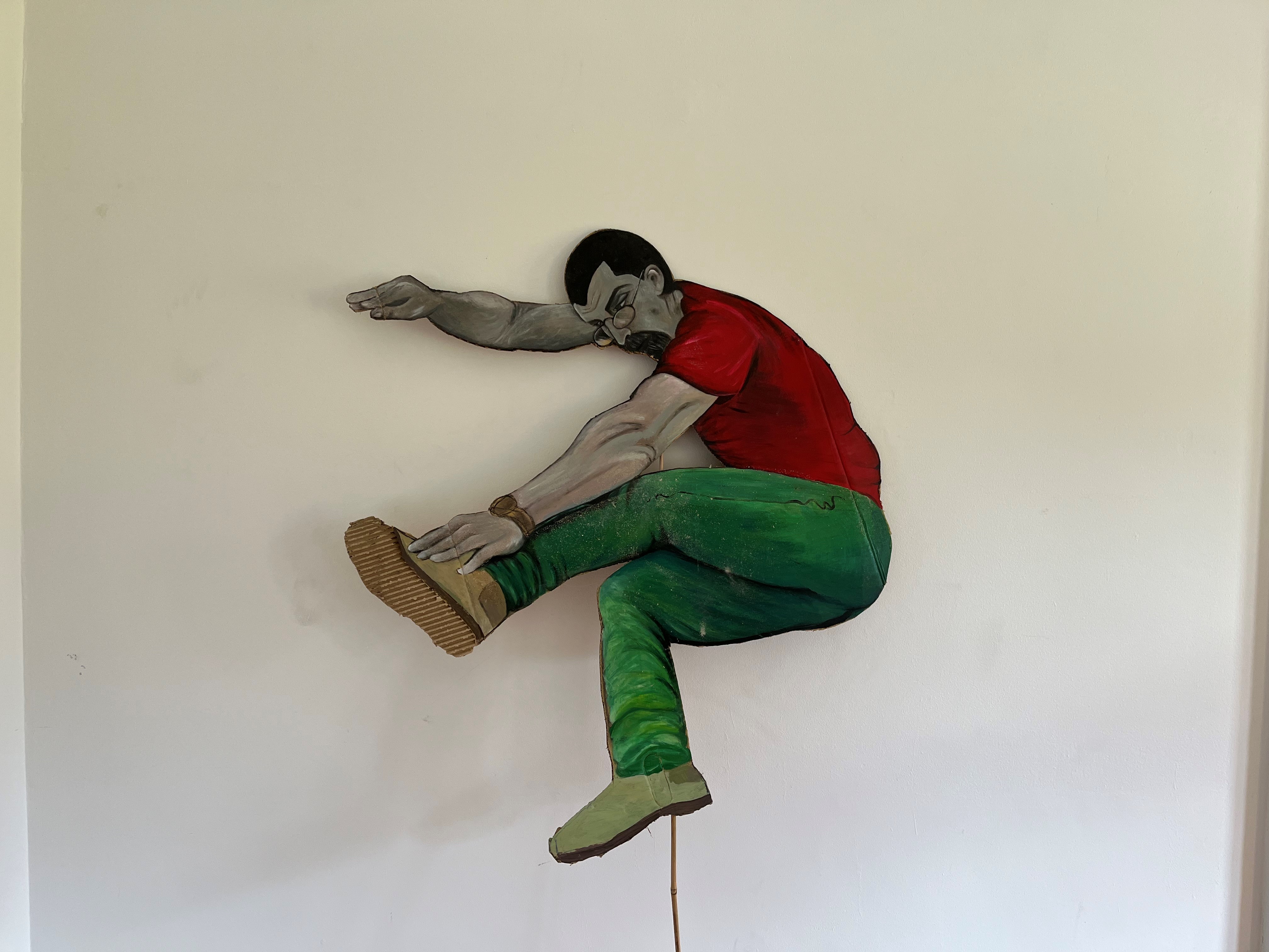 A man jumps mid air he wears green pants and a red top, his arm reaches his jumping leg an the other arm reaches out behind his ear